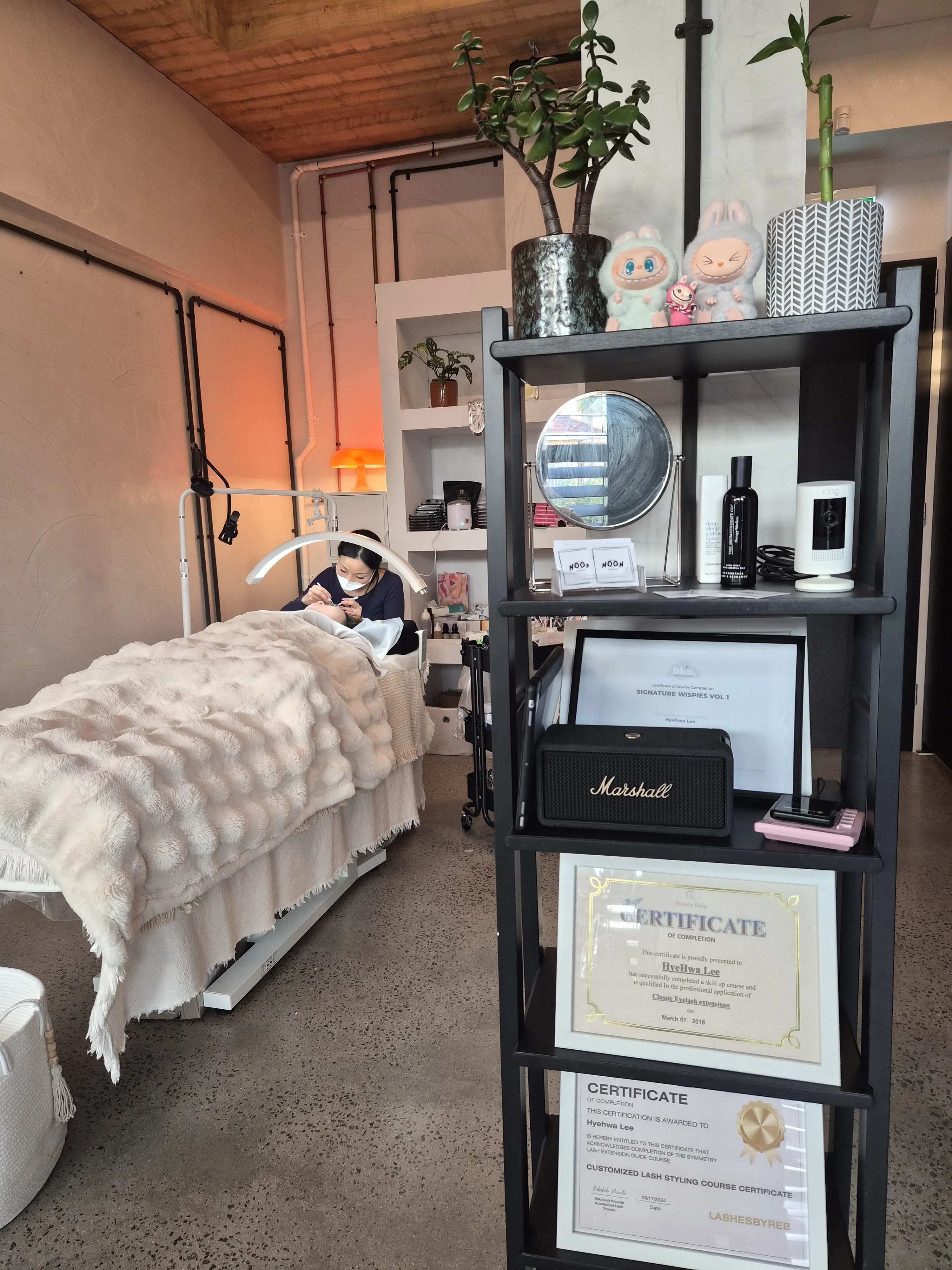 The Noon Lash & Co studio from the bed end — cream throw on the lash bed, black shelf ladder with framed training certificates and a Marshall speaker
